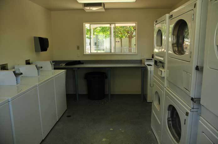 A photograph of the interior shared laundry room shows a stack of front-loading washers on one wall. On the opposite wall is a row of dryers. The back wall has a counter and a window.
