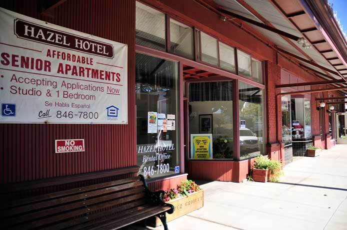 An exterior photo of the front door of the Hazel Hotel shows the large display-style windows. There's a bench to the side of the entrance. Two small planters are situated on either side of the entrance.