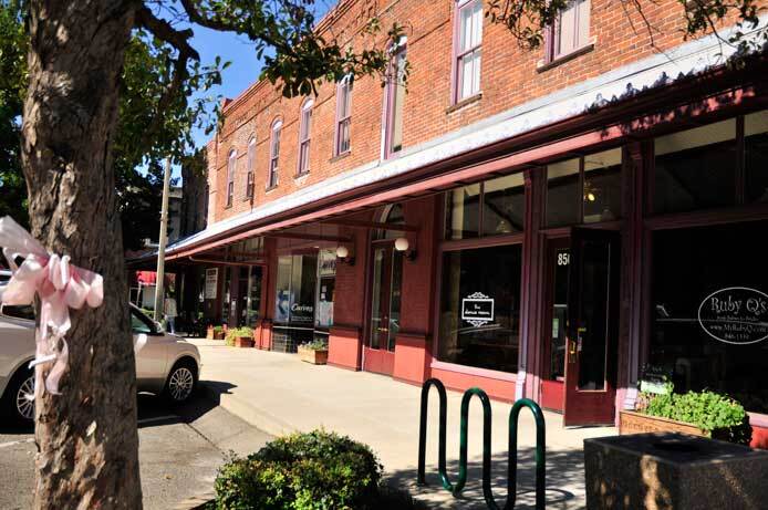 An exterior photo shows the front of the Hazel Hotel. There is a wide sidewalk that runs the length of the front of the building. An awning stretches over the front. Above the awning you can see the front second story of the building made of bricks. There are many windows in the front of the building. There's a pick rack on the sidewalk.