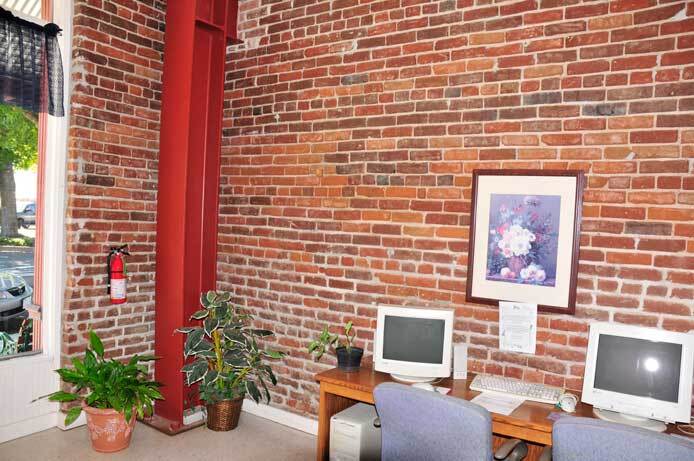 An interior photo shows the computer lab. It shows a room with tall ceilings and brick walls. There are two computers on a table with two chairs. Framed art is hanging above the computers on the table. There are two potted plants in the corner.