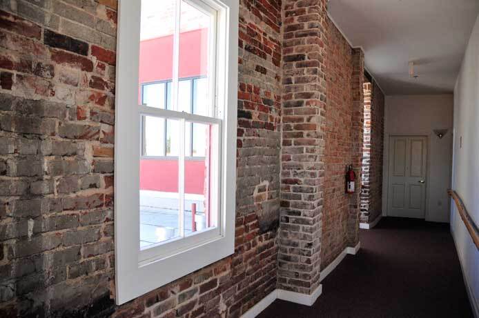 An interior photo shows a hallway. The exterior wall has a window and is made up of bricks. The opposite interior wall is painted white.